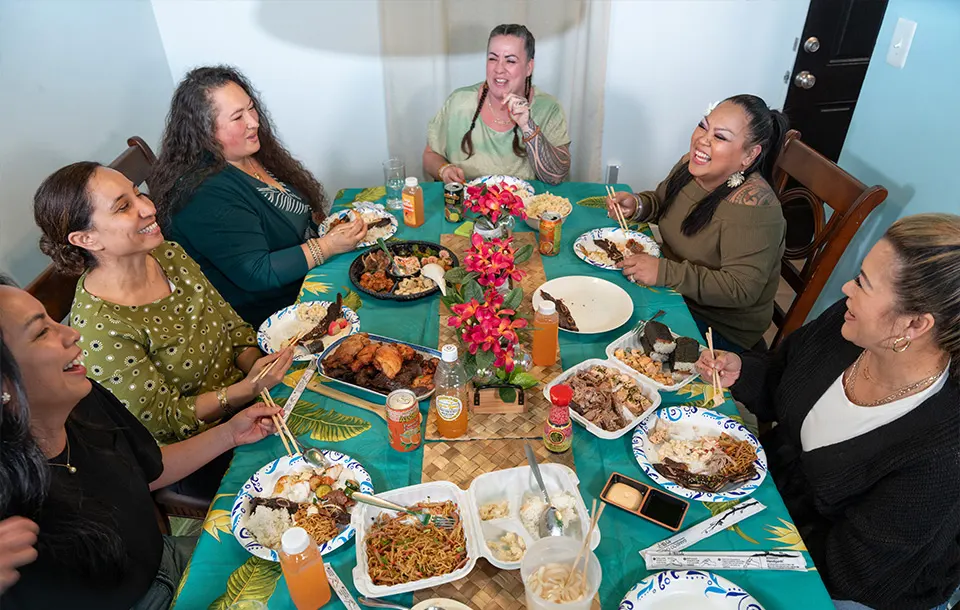 A joyful group enjoying a Hawaiian feast with diverse dishes from Pua's Plate Lunch catering, surrounded by a tropical-themed setting.