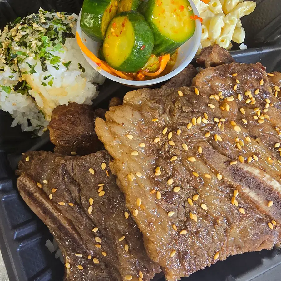 Plate of Hawaiian Kalbi Ribs with sesame seeds, cucumber salad, rice topped with furikake, and macaroni salad from Pua's Plate Lunch.