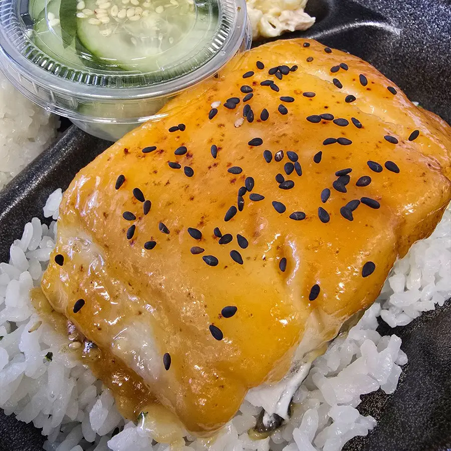 Misoyaki Butterfish over rice, served with a side of pickled cucumbers, representing Hawaiian cuisine.